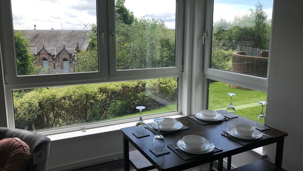 Comedor con una mesa dispuesta para cuatro personas, ventanas amplias con vistas a un jardín y un edificio.