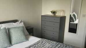 Una cama bien tendida con una manta a rayas y almohadas, un aparador gris con cajones, una mesita de noche pequeña y un espejo que refleja la cama y las almohadas.