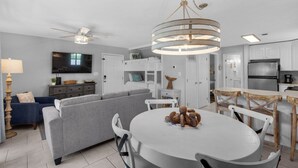 Uma sala de estar moderna com um sofá cinza, uma mesa de jantar com cadeiras, um ventilador de teto e uma cozinha com armários e eletrodomésticos brancos.