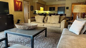A living room with a sofa, coffee table, and a TV. There is a kitchen in the background with cabinets and appliances. - Great Condo just 3 min walk to Lionshead Gondola. (Vail)