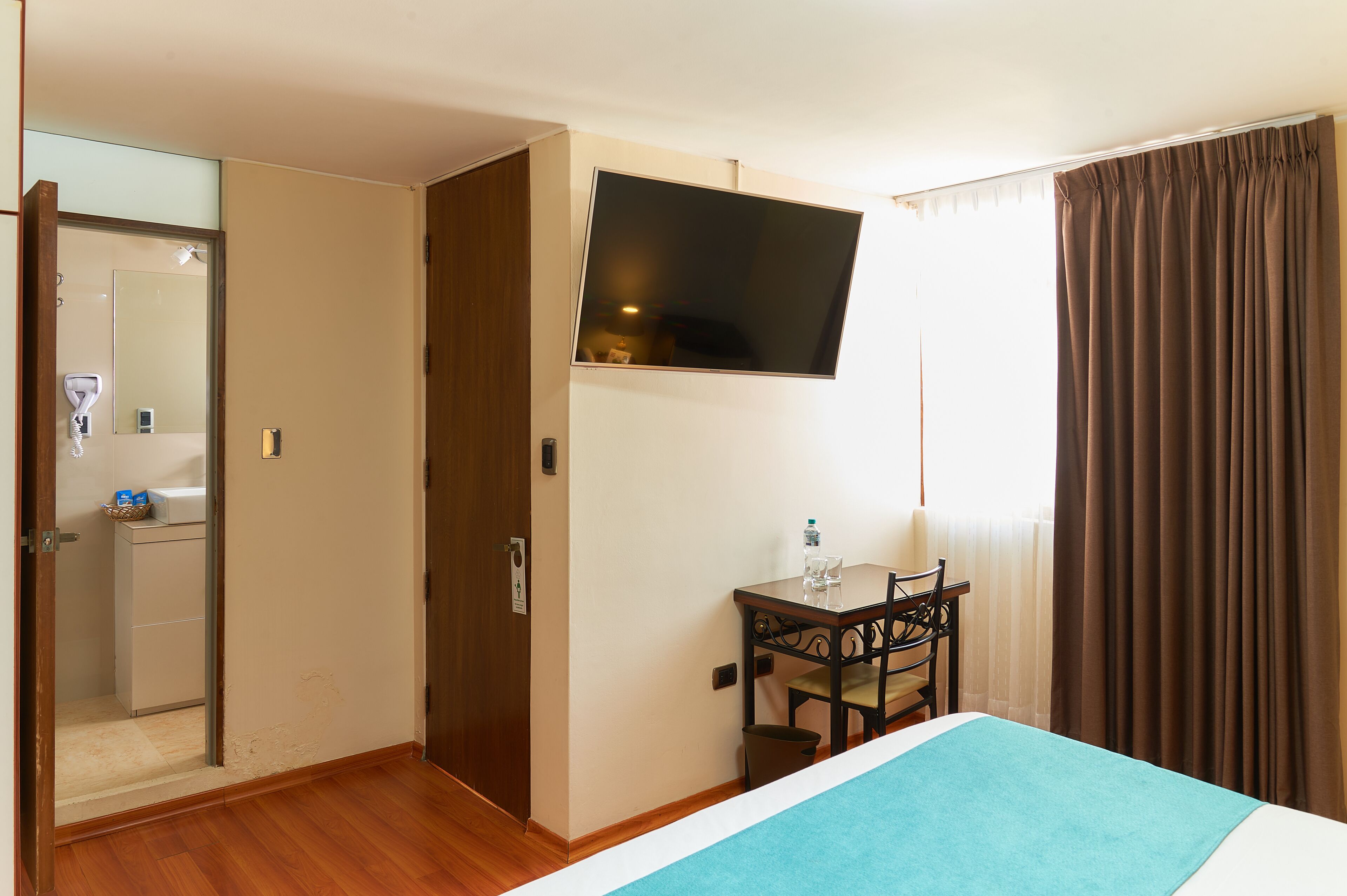 Habitación de hotel con cama, escritorio con silla, televisor montado en la pared y baño visible a través de una puerta abierta.