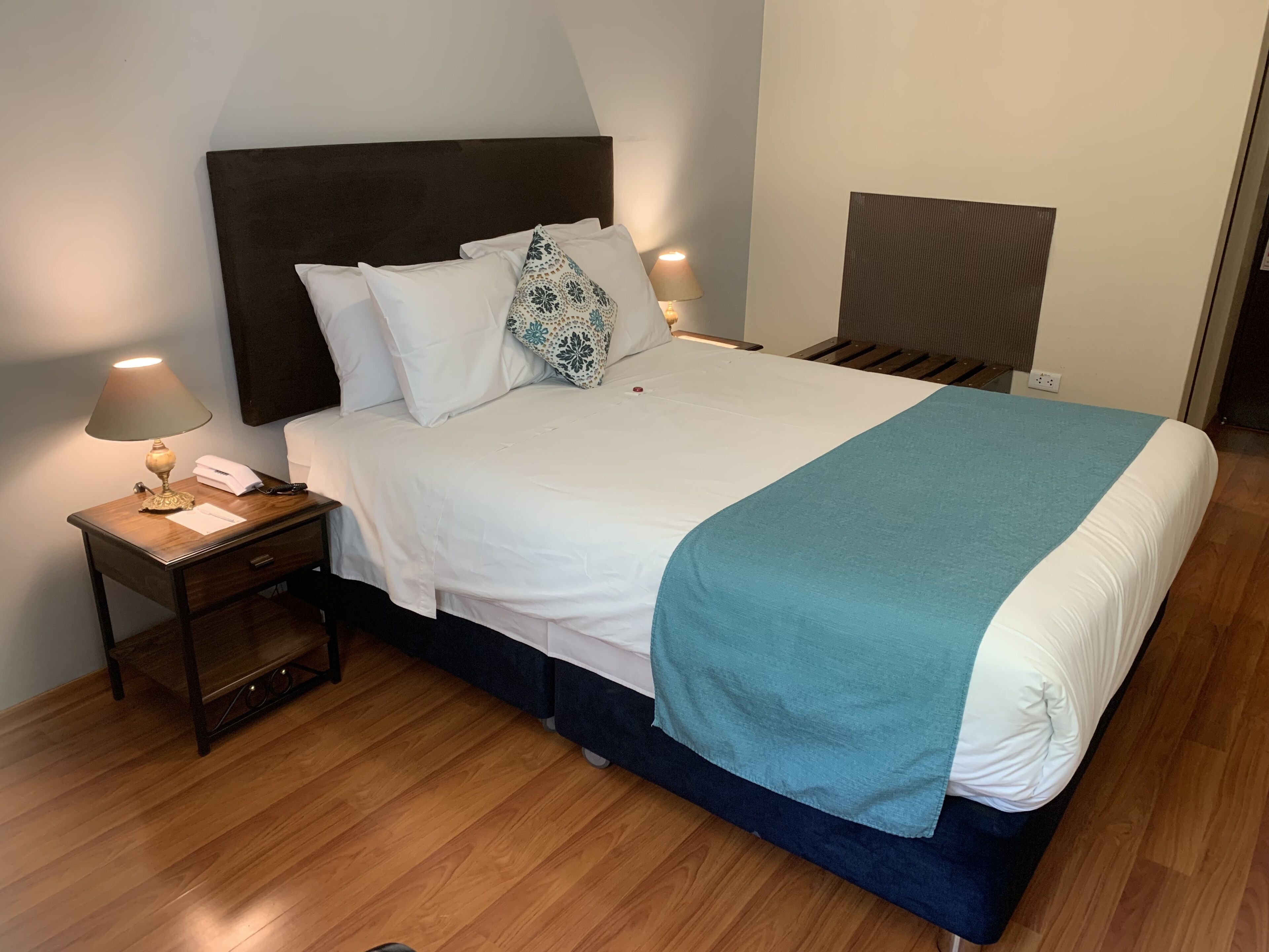 A neatly made bed with a white and teal bedspread, two bedside lamps, and a wooden nightstand with a lamp and a telephone.