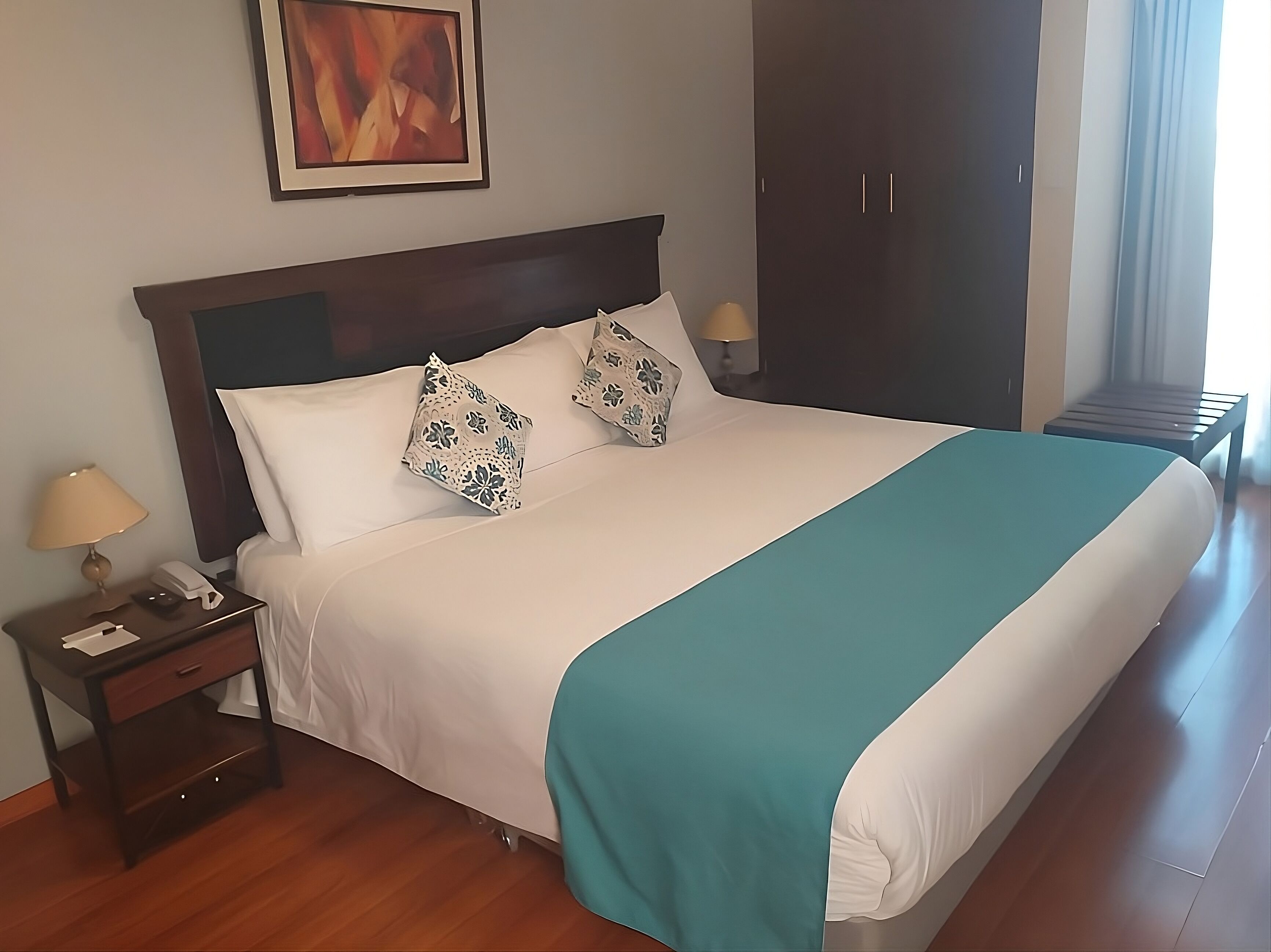 A neatly made bed with a teal bedspread, white pillows, and a wooden nightstand with a lamp and phone.