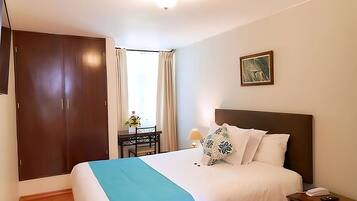 Una habitación de hotel con cama, escritorio, silla, armario y un cuadro en la pared.