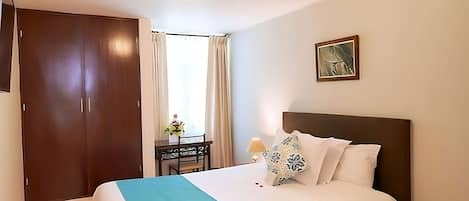 A hotel room with a bed, a desk with a chair, a wardrobe, and a painting on the wall.