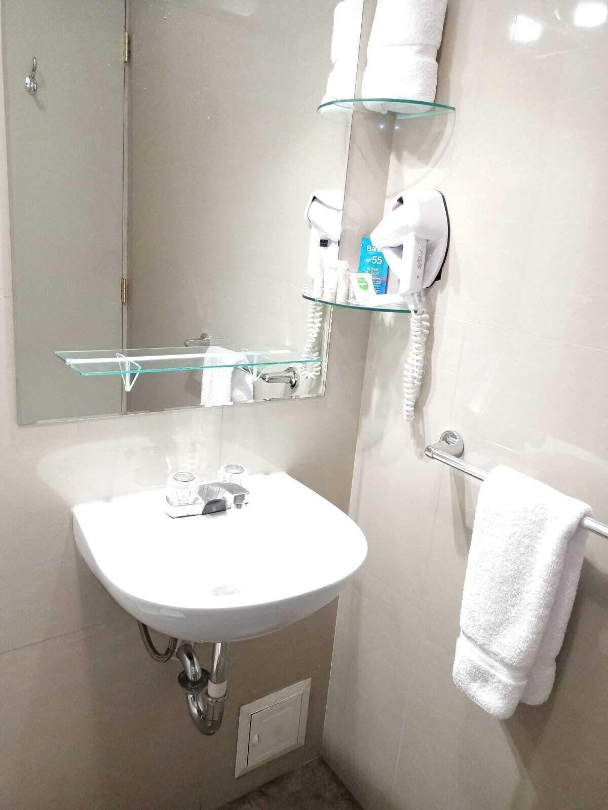 A bathroom with a white sink, a glass shelf above the sink, a towel rack with a white towel, and a hairdryer on the wall.