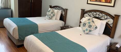 A hotel room with two beds, each with a teal blanket, white linens, and decorative pillows. There is a wooden wardrobe, a nightstand with a phone, and a framed abstract painting on the wall.
