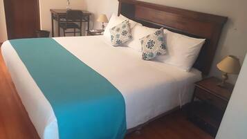 Una cama bien hecha con colcha azul verdosa y blanca, dos almohadas estampadas, un cabecero de madera, una mesita de noche con una lámpara y un escritorio pequeño con una silla y una botella.