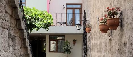 Un estrecho callejón con una escalera que lleva a un edificio con un depósito de agua en el techo. Hay plantas en macetas y muebles de exterior a lo largo de las paredes.