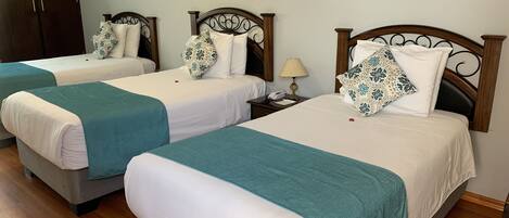 Habitación de hotel con dos camas individuales, cada una con colcha color turquesa y almohadas blancas, cabecero de madera y mesita de noche con lámpara.