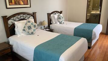 Habitación de hotel con dos camas, cada una con ropa de cama blanca y mantas turquesa. Se ve un mesita de noche con una lámpara y un cuadro en la pared.