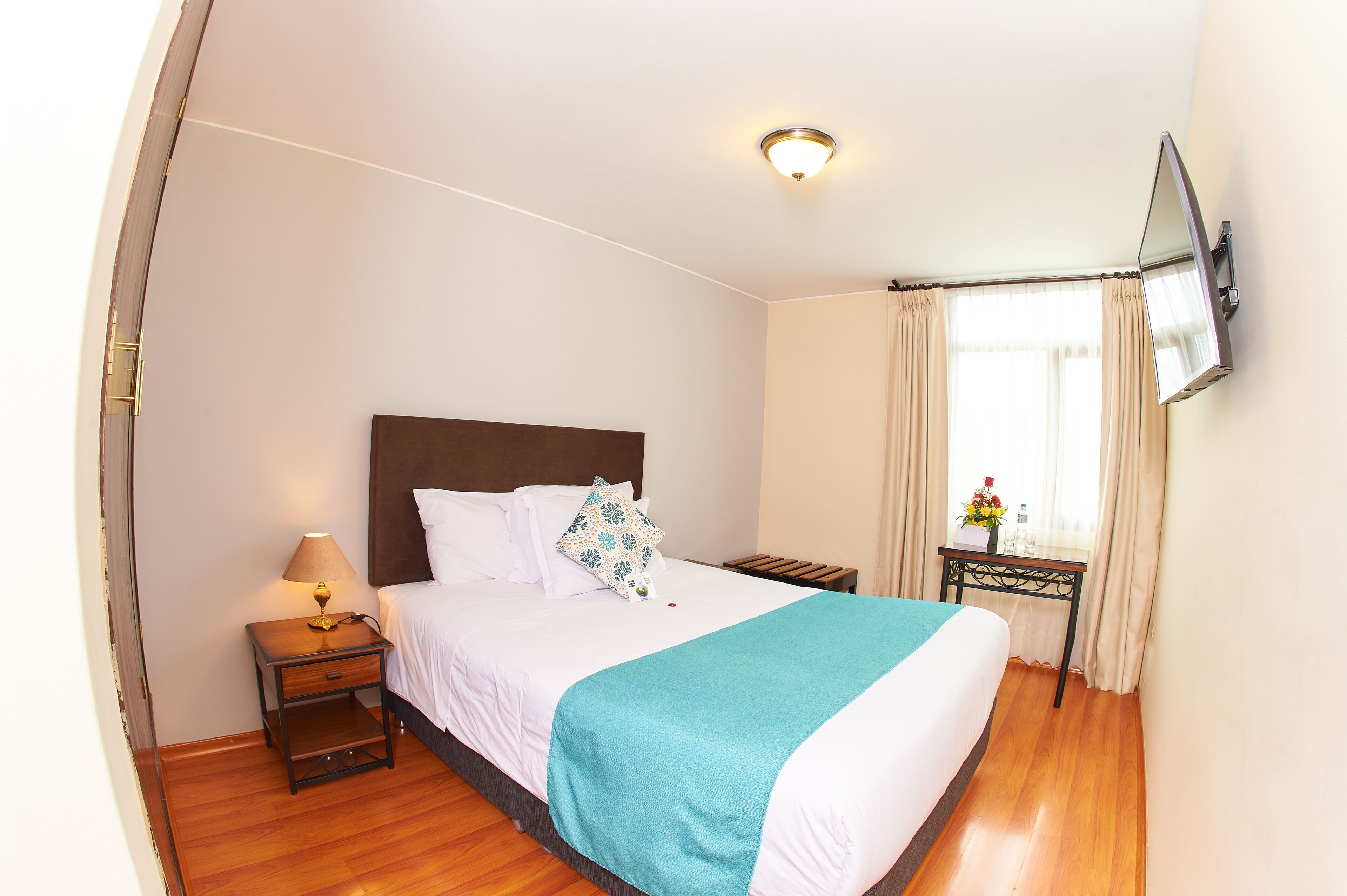 A neatly made bed with a headboard, a bedside table with a lamp, a TV mounted on the wall, and a window with curtains.