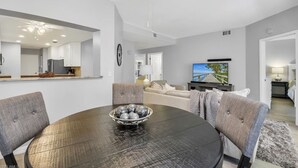 A modern living room with a round dining table, grey upholstered chairs, a large flat-screen TV, and a kitchen in the background. - Cozy Condo on Golf Course/Summer Rental/ Fort Myers  (Fort Myers)