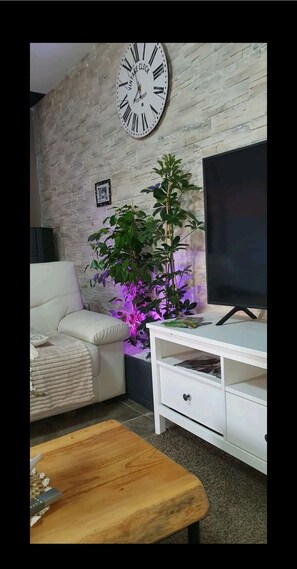 Una sala de estar con un sofá blanco, una mesa de centro de madera, un mueble blanco para televisor, un reloj grande y una planta en maceta.