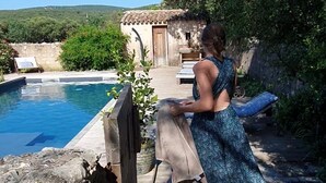 A person walking on a wooden deck towards a pool area with a stone wall and a small building in the background. - eco-lodge with natural swimming pool (PUECHABON)