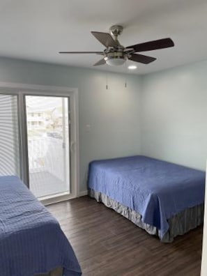 Un dormitorio con ventilador de techo, cama con cubrecama azul y vistas a través de un ventanal corredero.