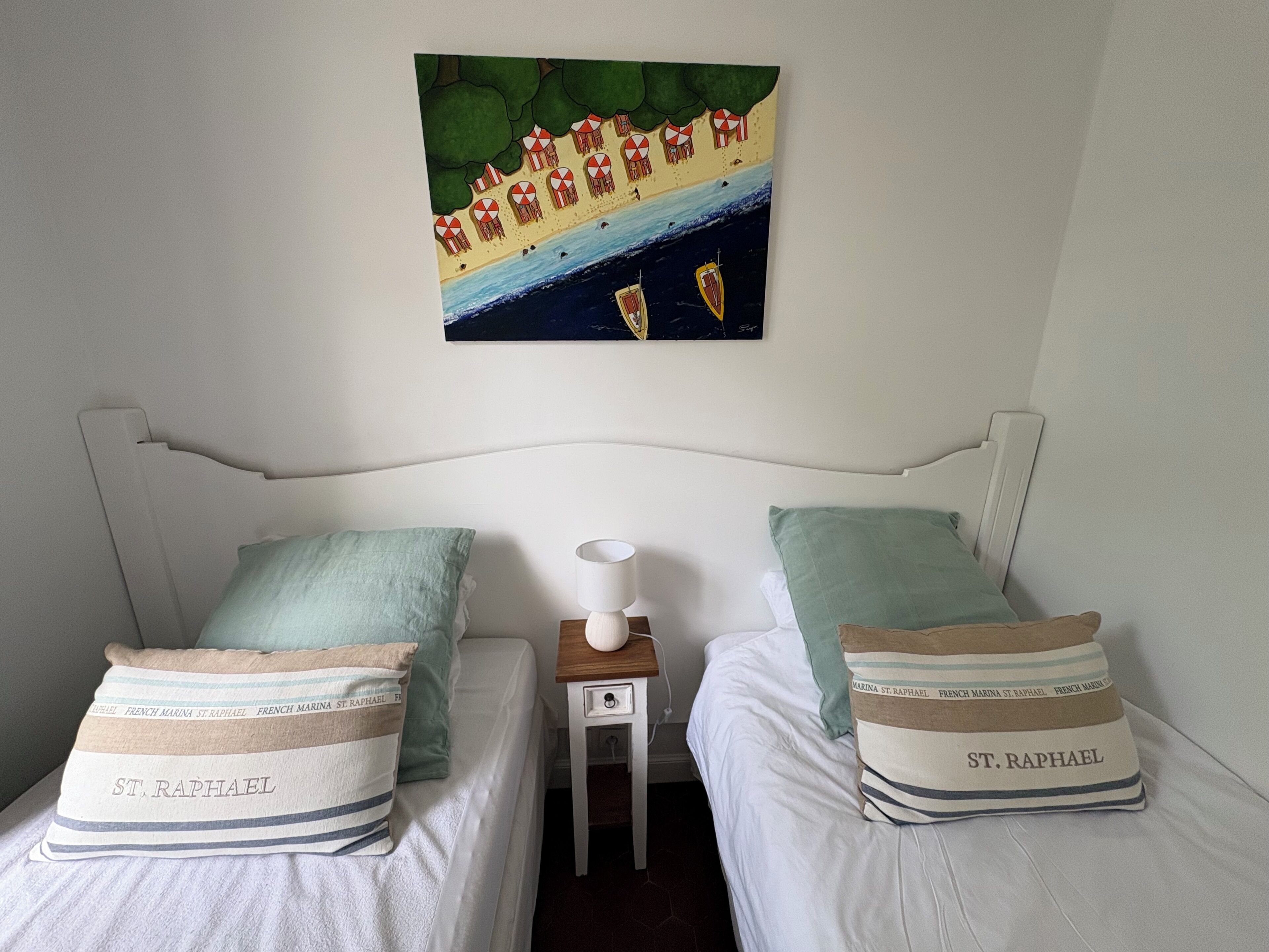 A bedroom with two beds, each with a pillow labeled 'St. Raphael'. A small bedside table with a lamp is between the beds.
