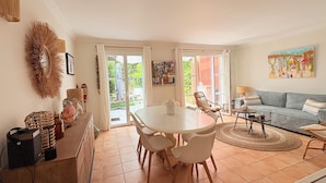 A spacious living area with a round dining table, white chairs, a sofa, and a coffee table. There is a large window with a view of greenery outside. - Villa 3 bed 2 bath 6 pers, south facing, Golfe St Tropez, air-conditioned, free wifi (Grimaud)