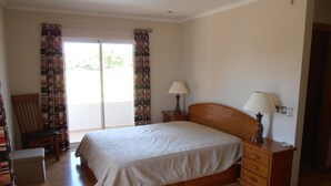 Un dormitorio con una cama de madera, una silla, una mesita de noche con lámparas y un ventanal corredizo al exterior.