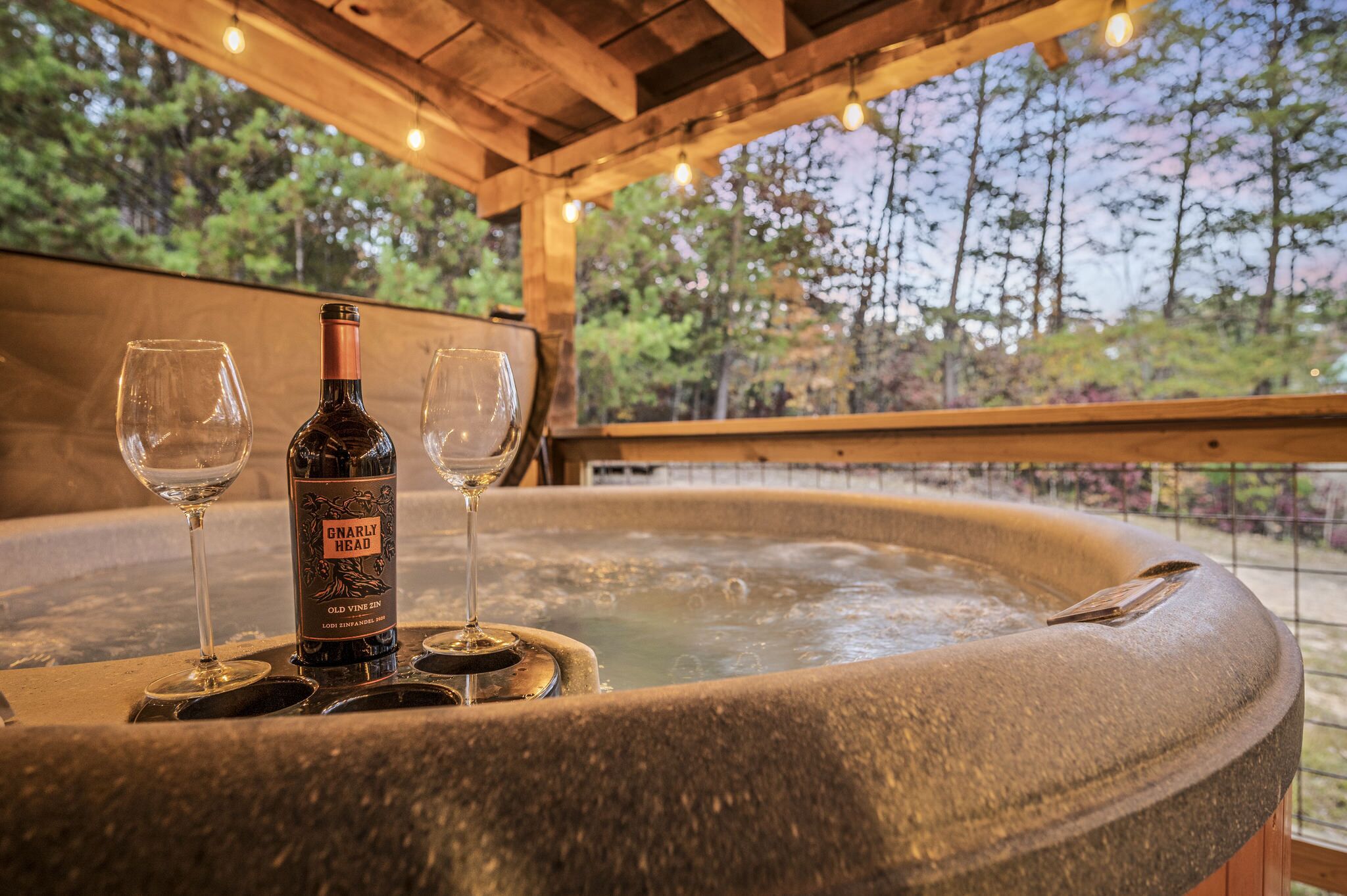 A hot tub with two wine glasses and a bottle of wine, surrounded by wooden beams and a view of trees.
