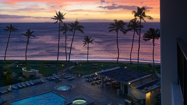 Un resort con piscina, sillas de descanso y palmeras frente al océano al atardecer.