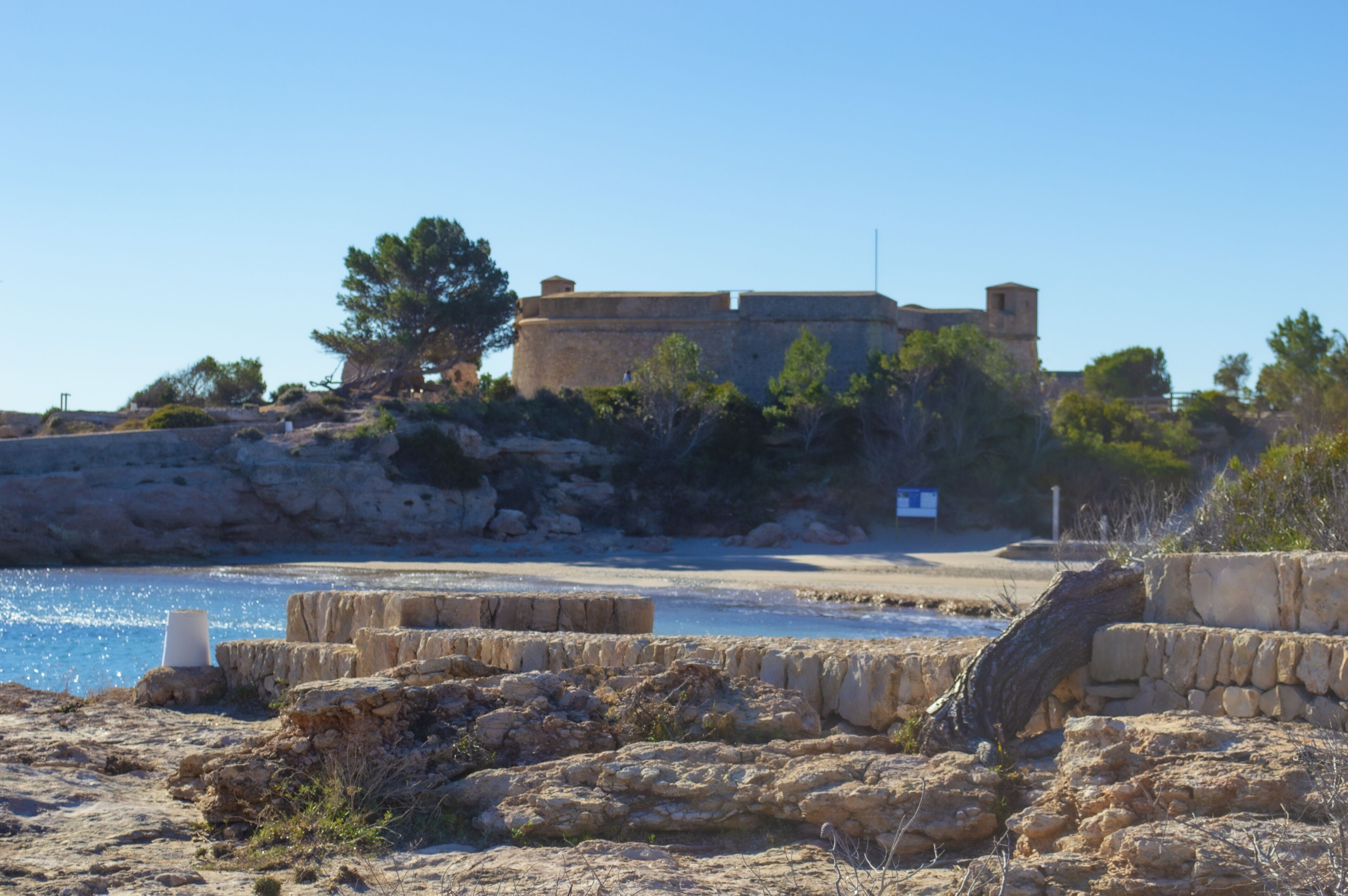 A coastal fortification with stone walls and a watchtower, surrounded by sparse vegetation and a clear sky.