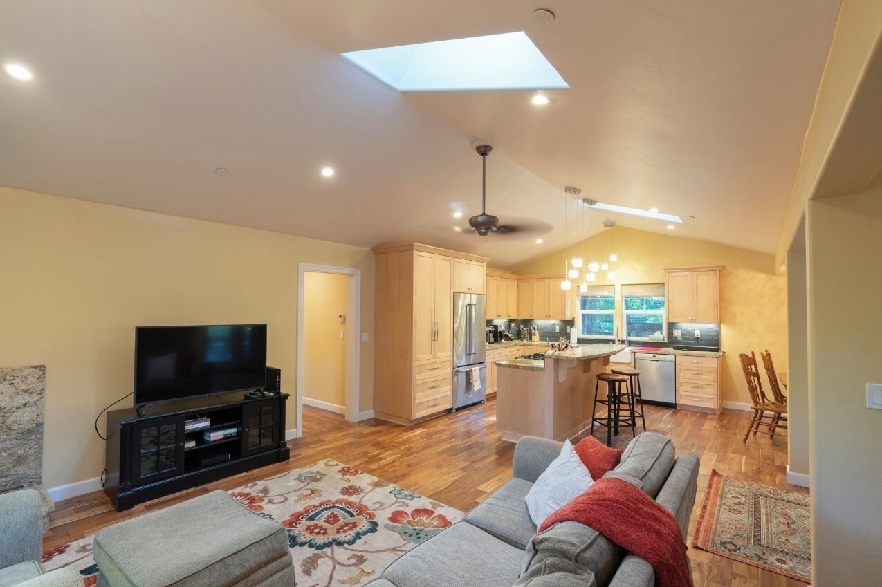 A spacious living area with a flat-screen TV, a sofa, and a coffee table. There is a kitchen with wooden cabinets and a dining area with a bar stool.