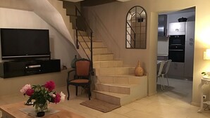 A living room with a TV, a wooden table with flowers, a chair, a staircase, and a kitchen area. - Warm, air-conditioned, 2-bedroom house 3 km from La Ciotat beaches. (Ceyreste)