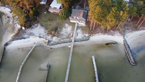 Vista aérea de una propiedad costera con playa de arena, un muelle de madera y una casa con terraza.