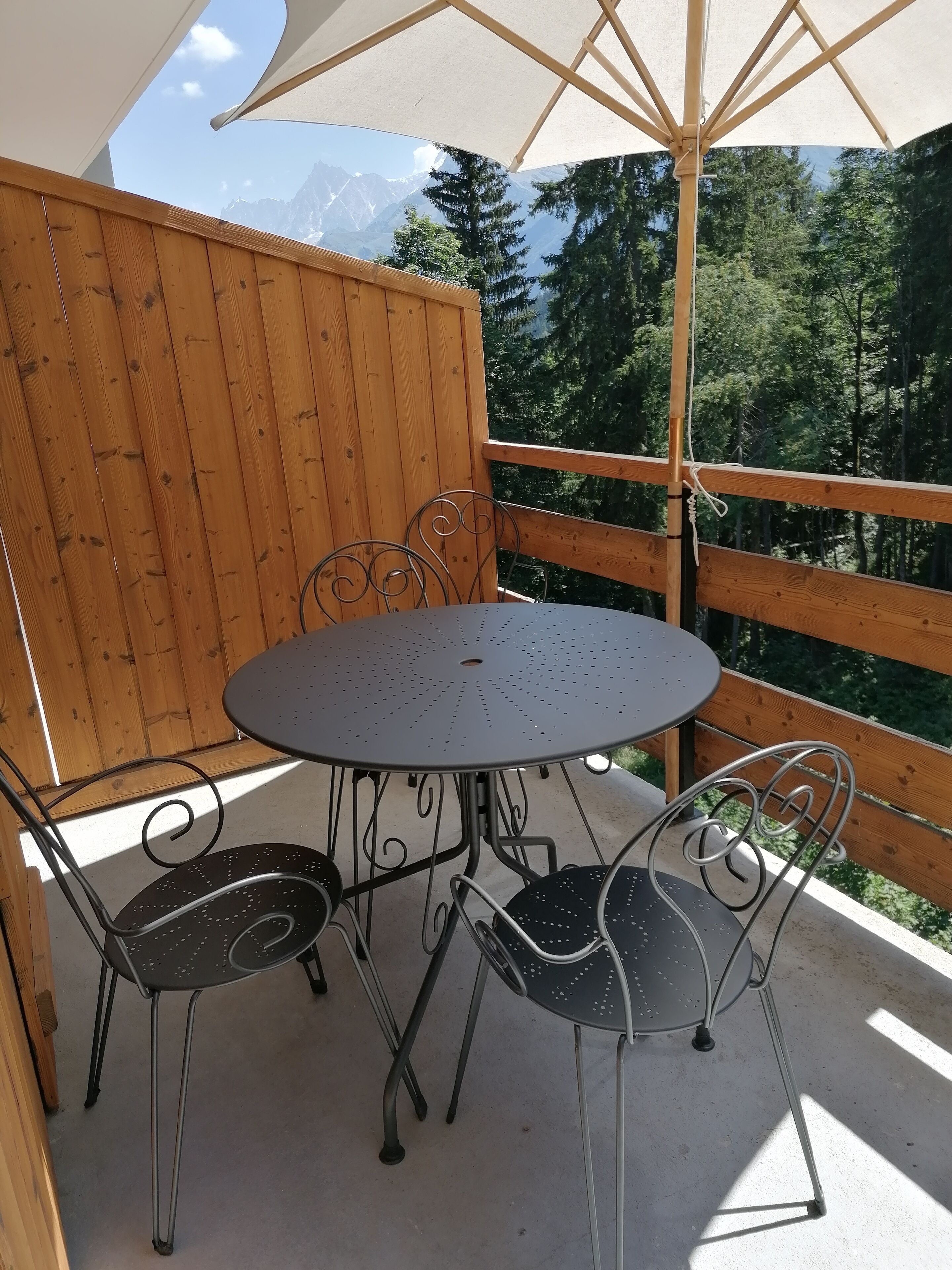 Una terraza de madera con una mesa redonda de metal y sillas, un gran parasol y vistas a montañas y árboles.