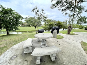 A concrete outdoor seating area with a table and benches, surrounded by trees and a body of water.