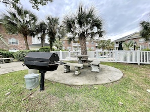 A barbecue grill, picnic table, and chairs in a grassy outdoor area with palm trees and a white fence.