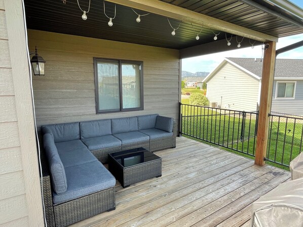 Eine überdachte Terrasse mit einem grauen Ecksofa und einem kleinen schwarzen Tisch, Holzboden sowie Blick auf ein Haus und Berge im Hintergrund.