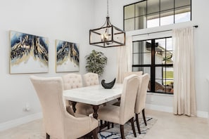 A dining room with a marble table, six upholstered chairs, a chandelier, two paintings, a potted plant, and a view through a large window.