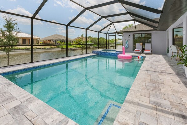 A swimming pool with a clear blue water, surrounded by a covered patio area with lounge chairs and a view of a lake and houses in the background.