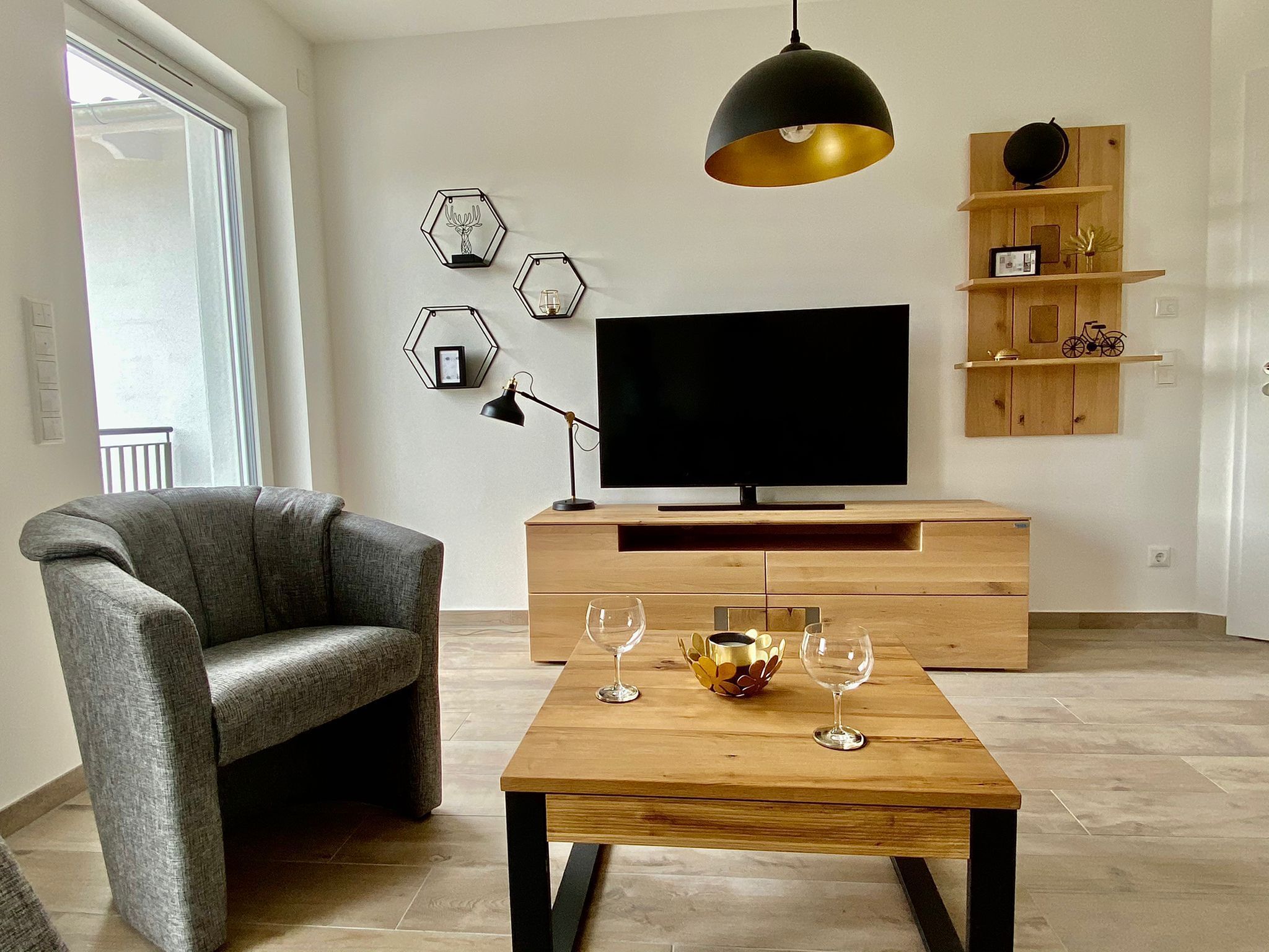 A modern living room with a grey armchair, a wooden coffee table with wine glasses, a TV on a wooden stand, and hexagonal wall art.