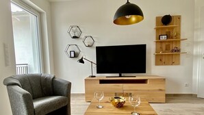 Una sala moderna con un sillón gris, una mesa de centro de madera con copas de vino, un televisor sobre un mueble de madera y decoración mural hexagonal.