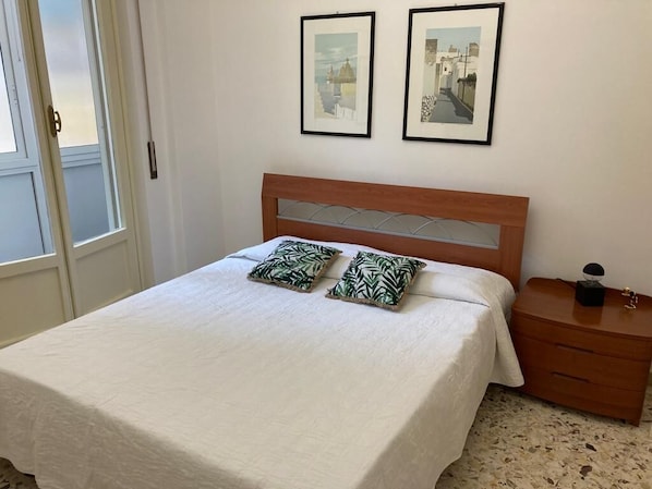 Un dormitorio con una cama, dos cuadros en la pared, una mesita de noche de madera y una puerta.