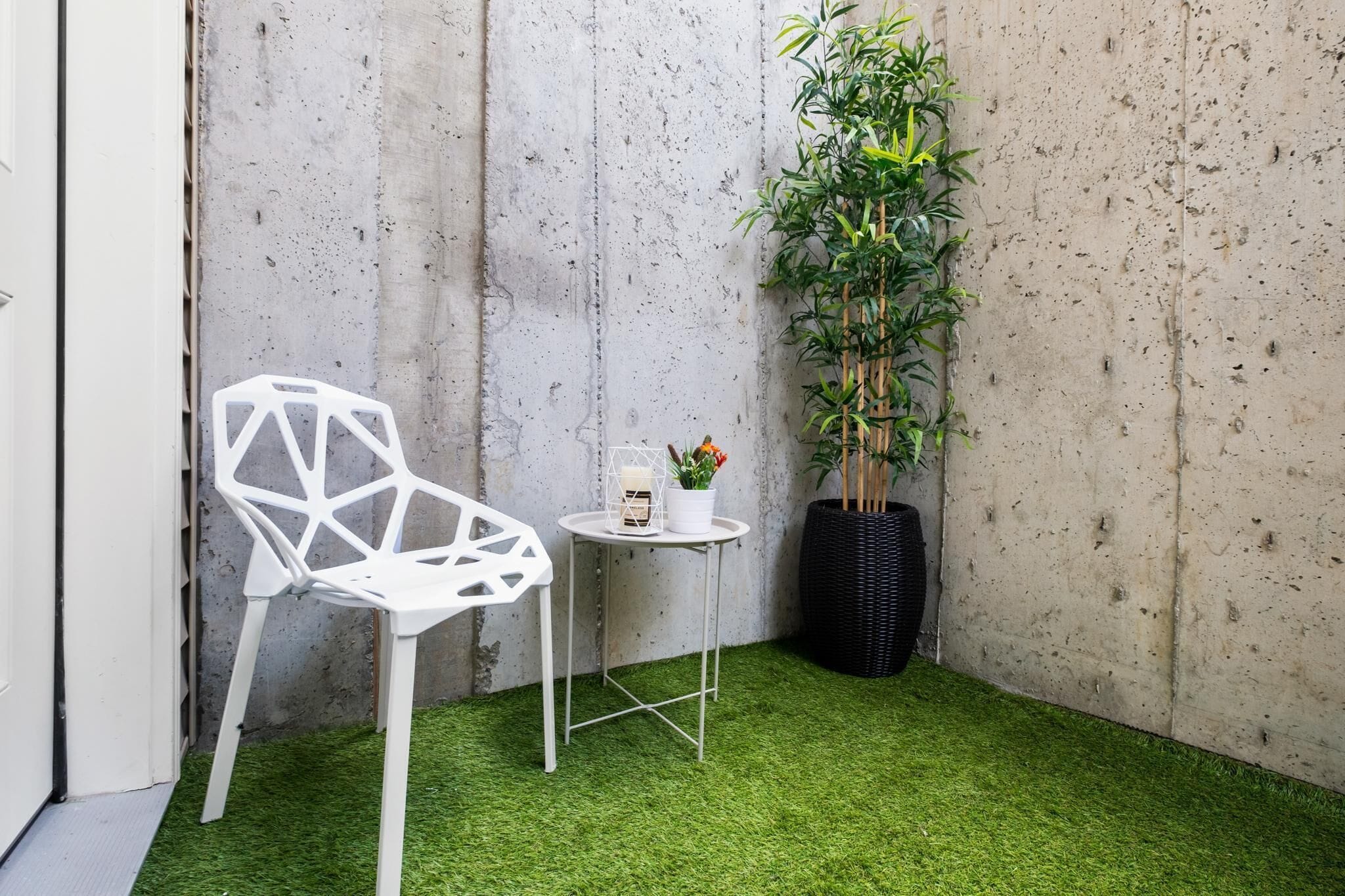 Un espacio pequeño y cerrado con una silla blanca, una mesita con una vela y flores, y una planta en maceta junto a una pared de concreto.