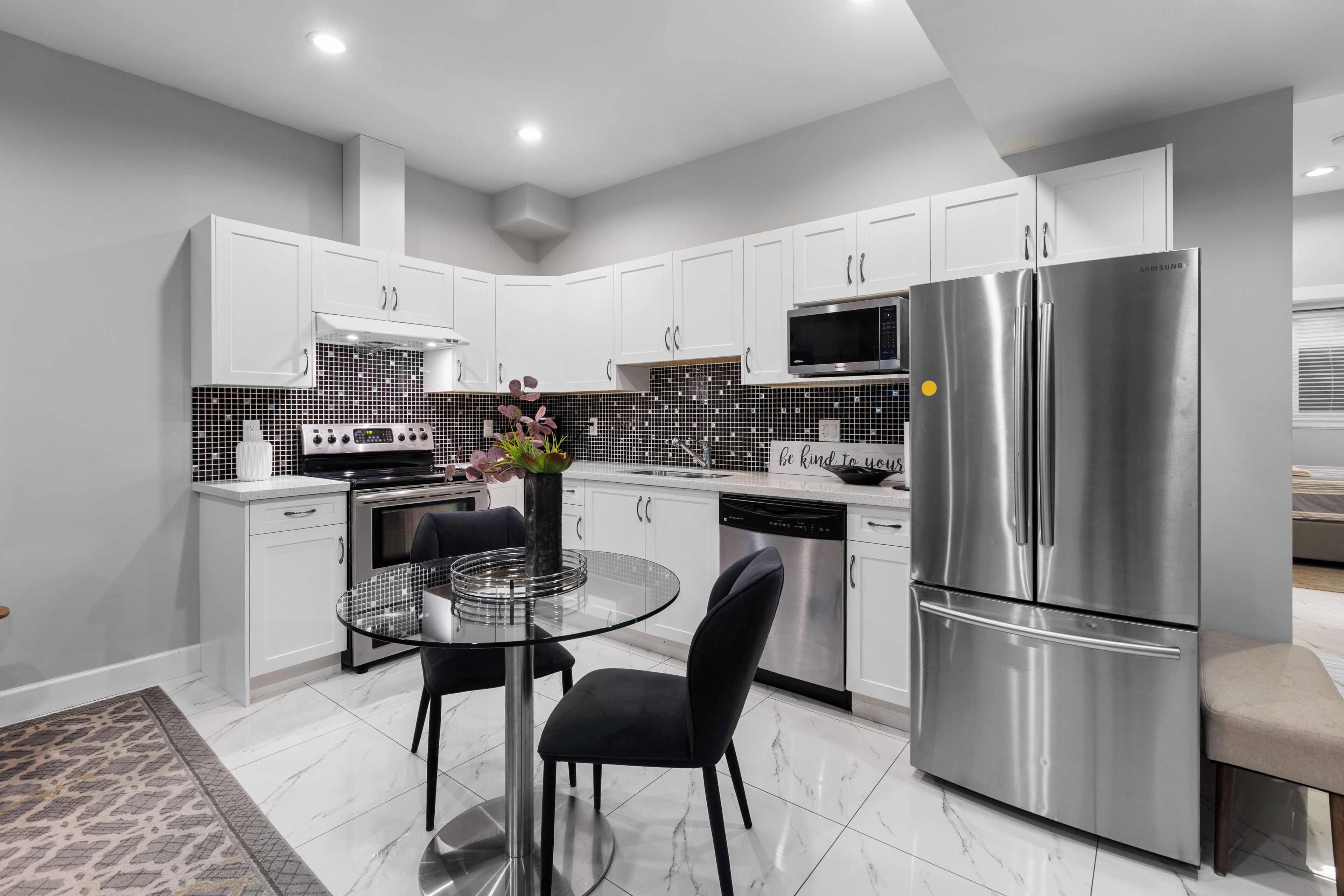 A modern kitchen with white cabinets, stainless steel appliances, a glass dining table, and a black chair.