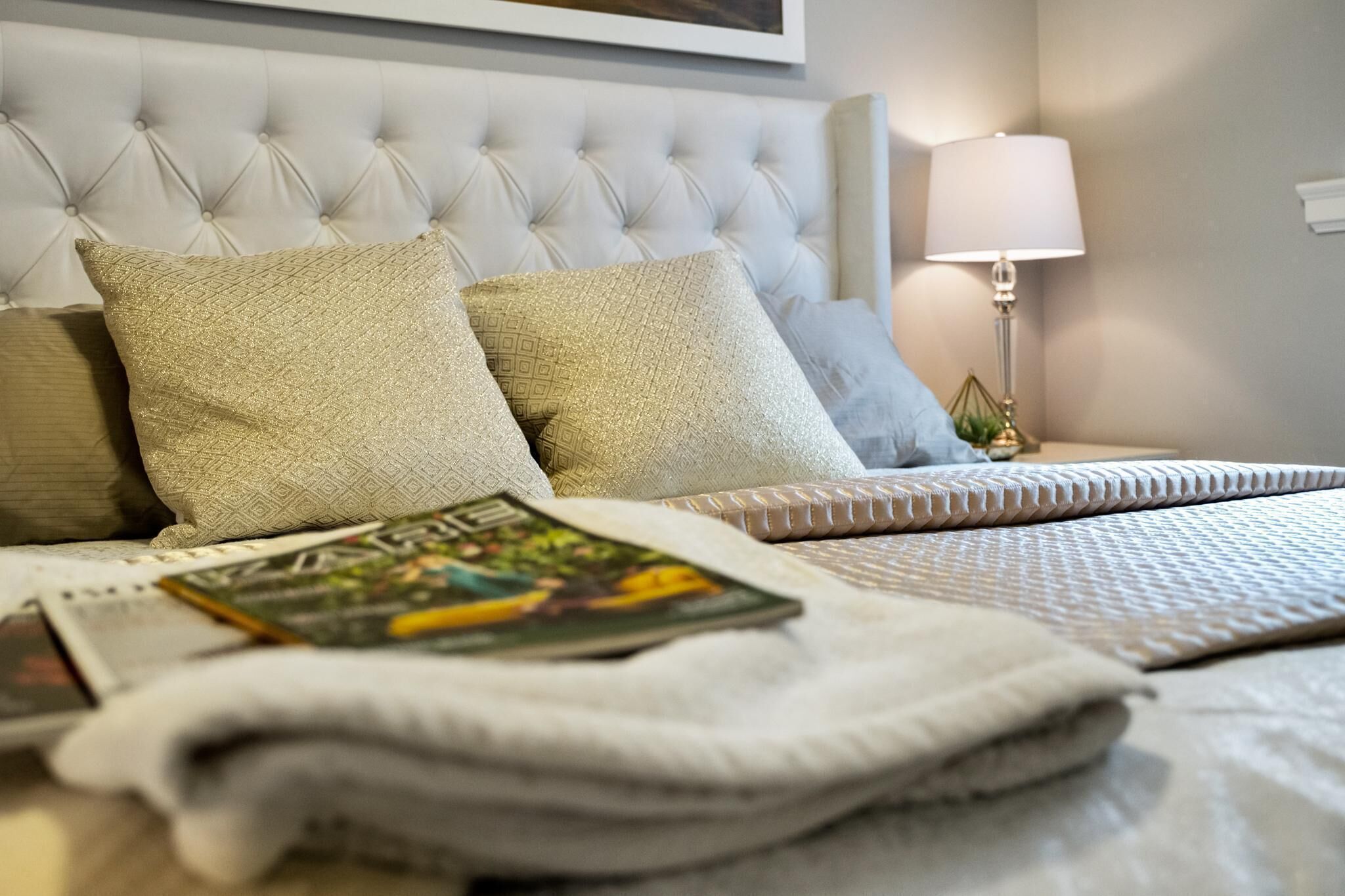 Een net opgemaakt bed met een gestoffeerd hoofdbord, een lamp en een tijdschrift op het bed.