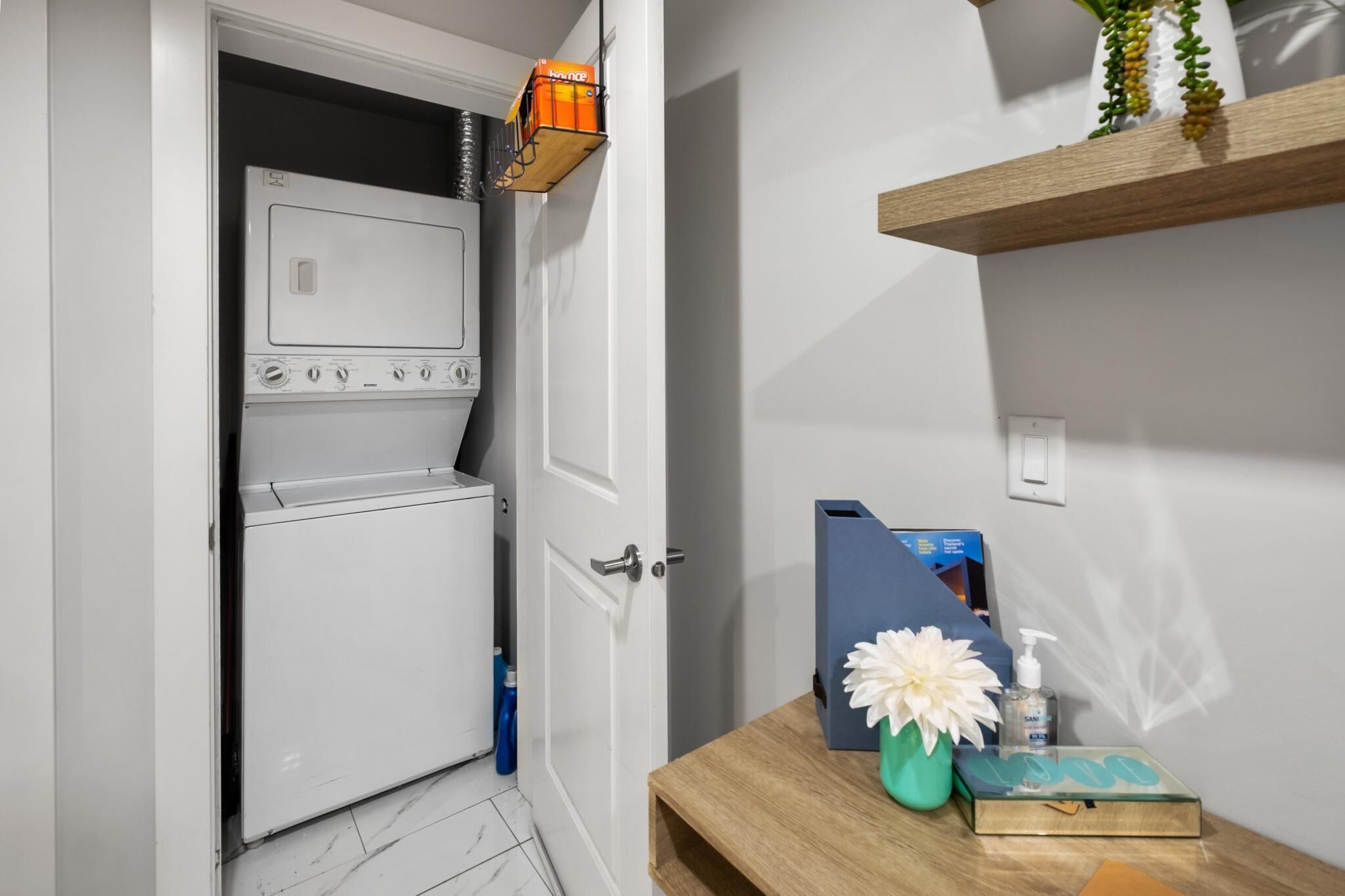 A laundry room with a white washing machine and dryer, a door with a handle, and a small table with a flower vase and a blue box.
