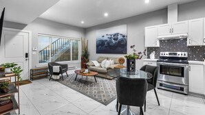 Ein modernes Wohnzimmer mit Sofa, Couchtisch und Sesseln, eine Küche mit weißen Schränken und einem Herd sowie eine Treppe, die zu einer weiteren Ebene führt.