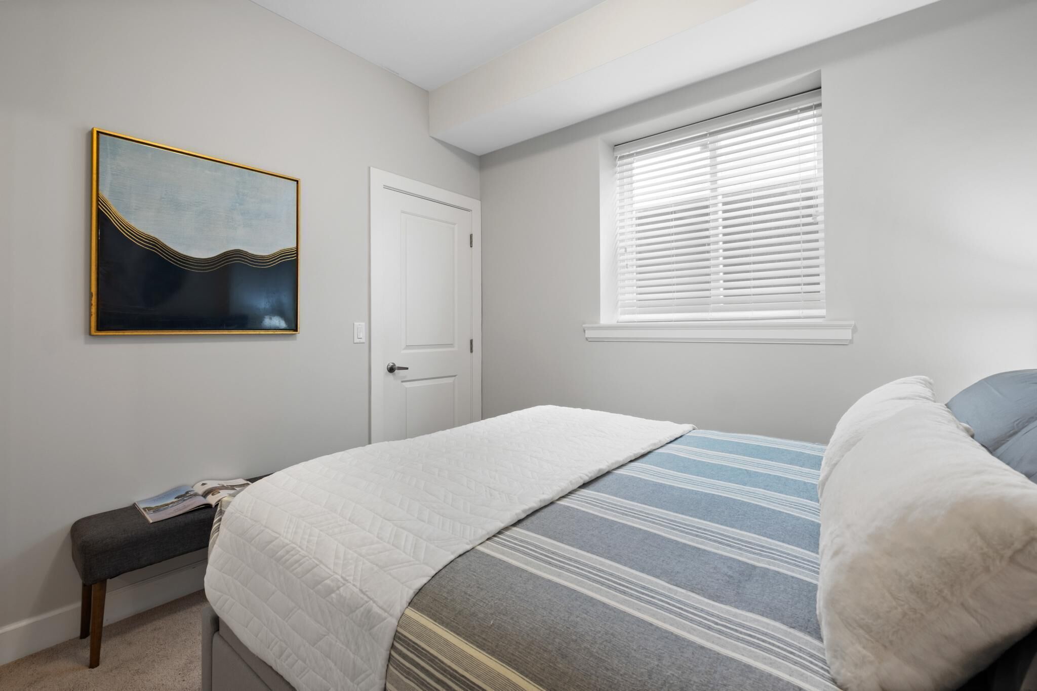 A bedroom with a bed, a window with blinds, a door, and a wall-mounted artwork.