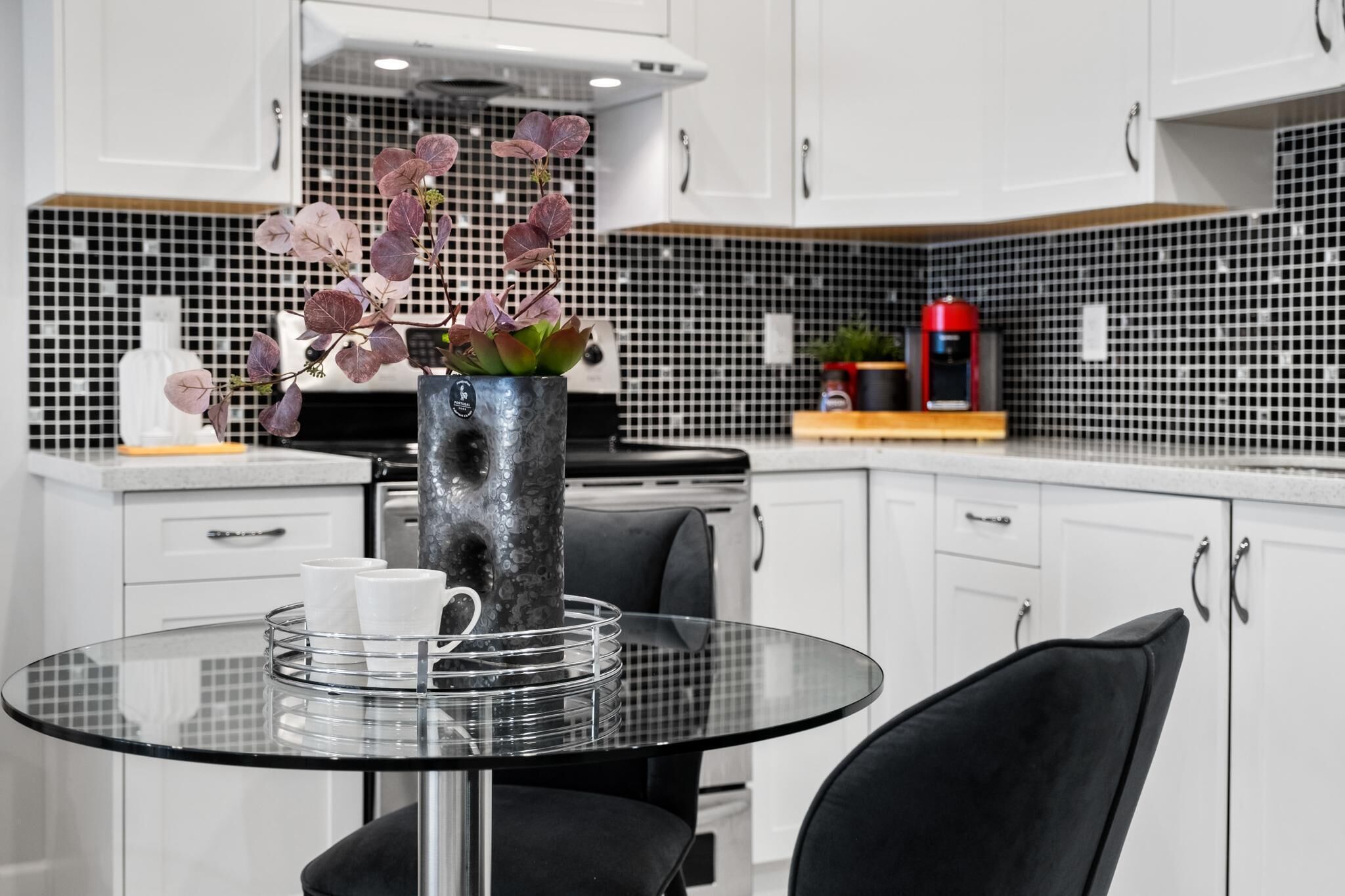 Una cocina moderna con armarios blancos, salpicadero de azulejos blanco y negro, una mesa de centro de vidrio y un jarrón con flores moradas.