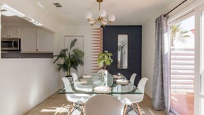 A modern dining area with a glass table, white chairs, a potted plant, and a dark accent wall. - The Yorba Oasis w/Pool (Palm Springs)