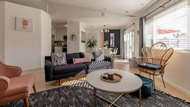 A modern living room with a black sofa, a round coffee table, and a wicker chair. A dining area is visible in the background with a table and chairs. - The Yorba Oasis w/Pool (Palm Springs)