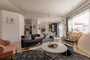 A modern living room with a black sofa, a round coffee table, and a wicker chair. A dining area is visible in the background with a table and chairs. - The Yorba Oasis w/Pool (Palm Springs)