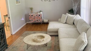 A living room with a beige sectional sofa, a round coffee table, a patterned rug, a decorative wall piece, and a small side table with plants. - Cush Jax Baymeadows Ranch, Close to Everything! (Jacksonville)
