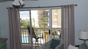 A living room with a sofa, a table, and a chair. There is a sliding glass door leading to a balcony with a pool and a view of buildings. - Oceanfront condo with beach view – 2 pools (Galveston)